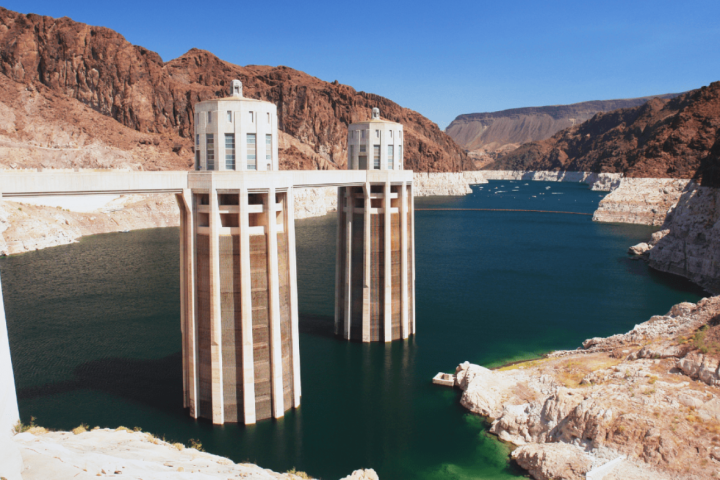 Two concrete intake towers at a dam with a reservoir, surrounded by rocky terrain and blue sky.